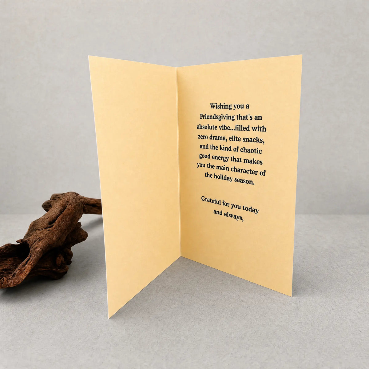 A folded greeting card standing open on a light gray surface, with a piece of driftwood to the left. The card is a warm pale yellow color with black printed text on the inside.