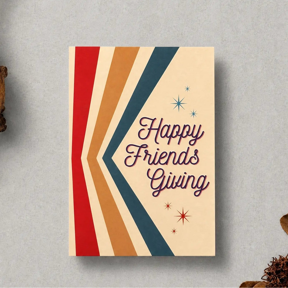 A greeting card with retro chevron stripes in red, orange, beige, and navy, featuring the text 'Happy Friends Giving' in purple script. The card is styled on a textured gray background with dried botanical props.
