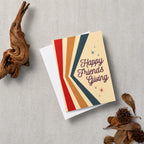 A greeting card with the words 'Happy Friends Giving' in dark script, layered over a white envelope. The card features retro diagonal stripes in red, orange, tan, cream, and dark teal, accented with small star details.