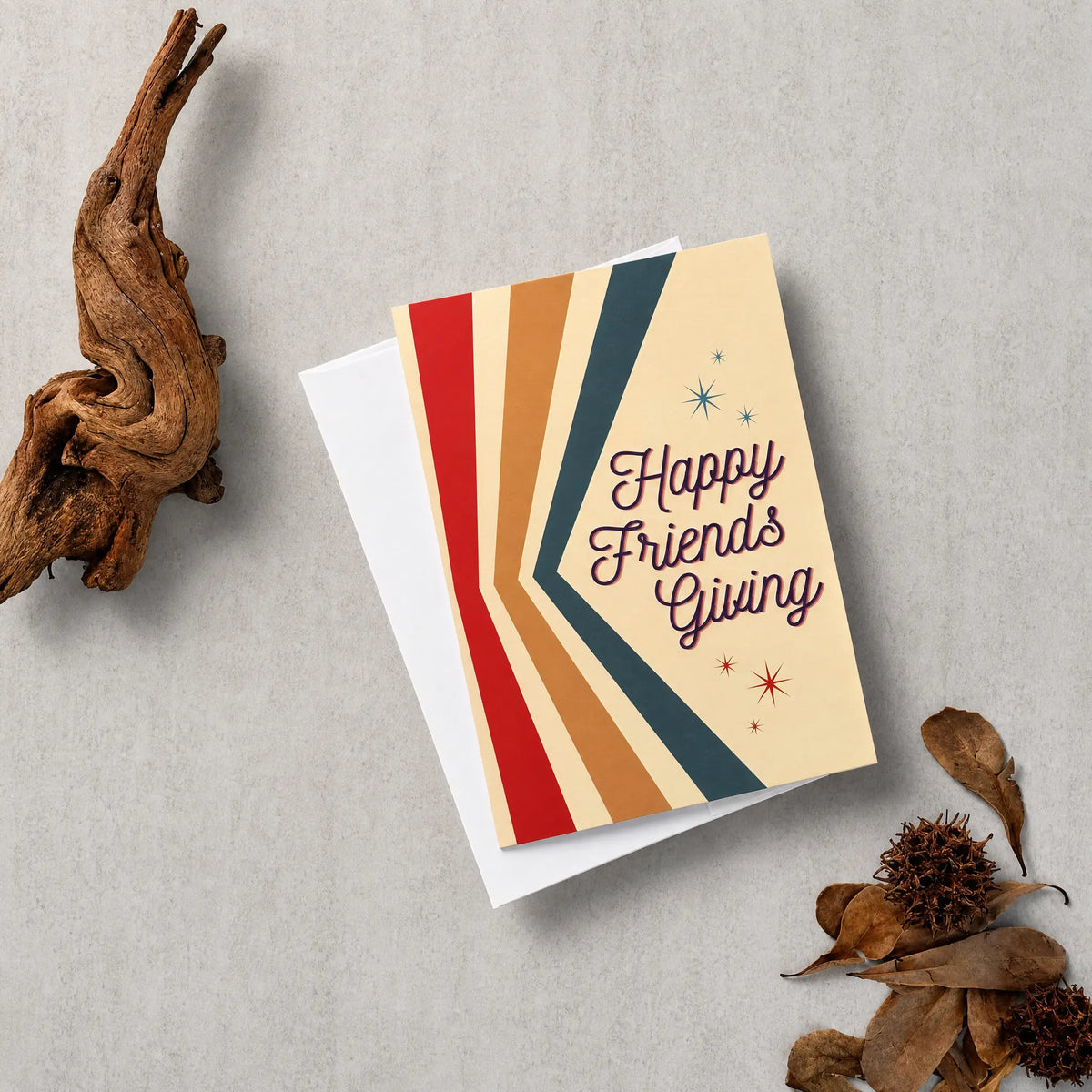 A greeting card with the words 'Happy Friends Giving' in dark script, layered over a white envelope. The card features retro diagonal stripes in red, orange, tan, cream, and dark teal, accented with small star details.