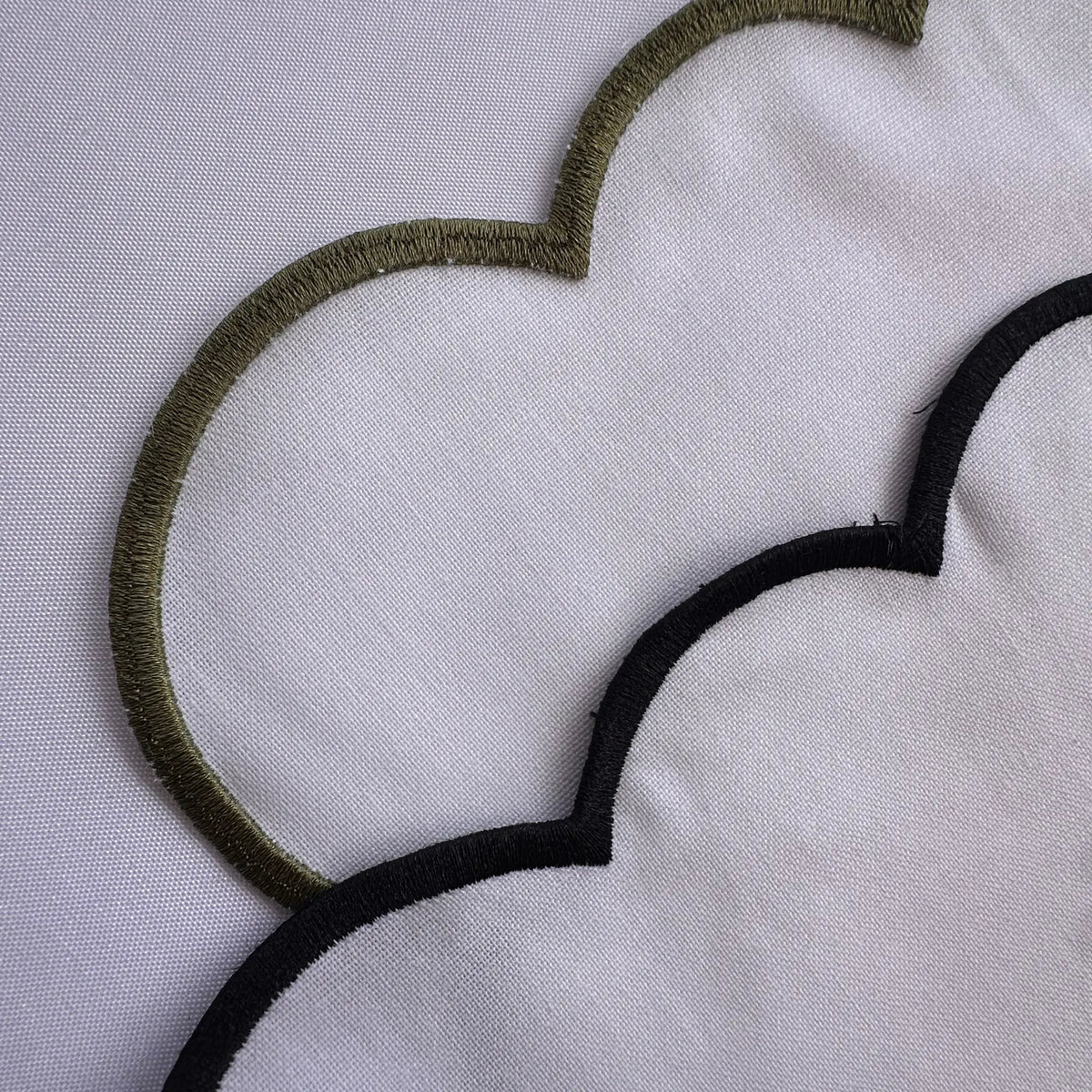 Close-up of a white fabric surface with embroidered scalloped outlines, showing an olive green outline at the top left and a black outline at the bottom right.