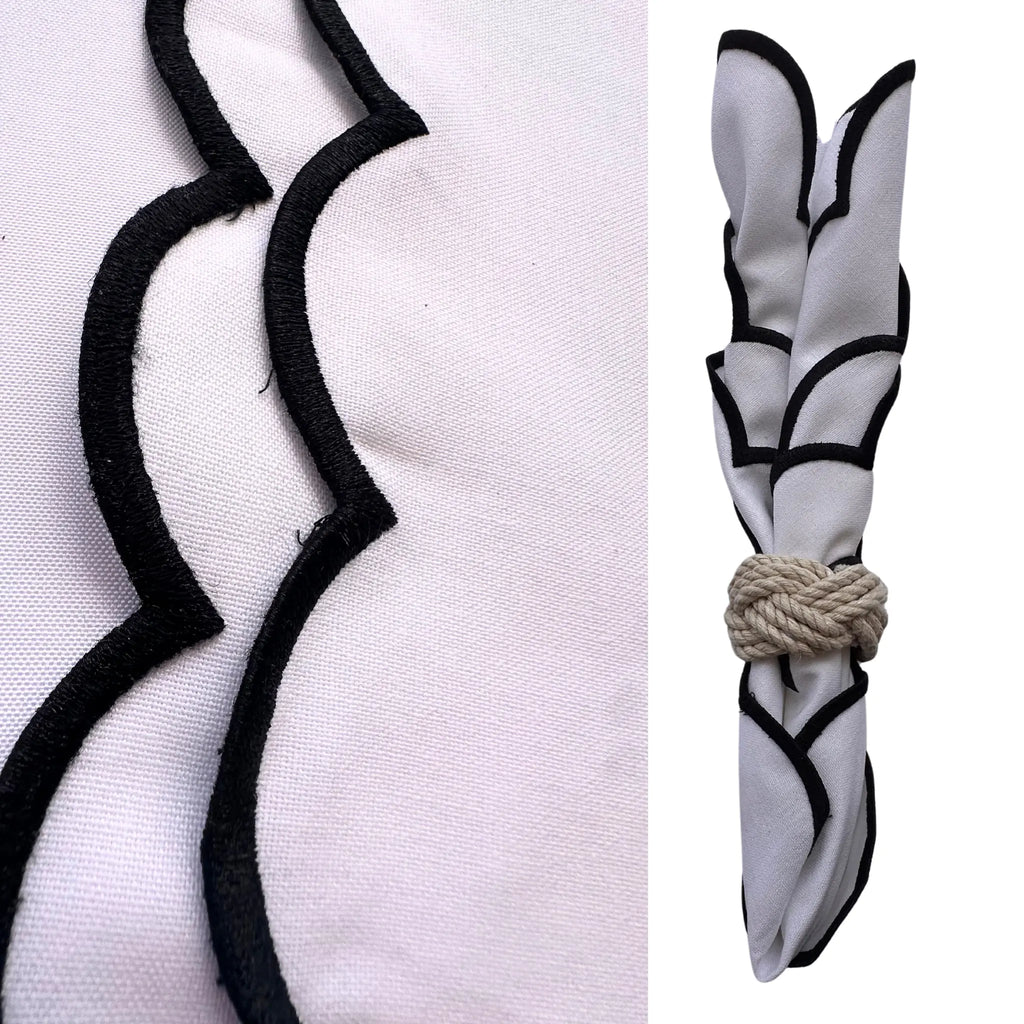 A white fabric napkin with black scalloped trim, shown in a close-up detail view on the left and folded with a rope tie on the right, against a white background.