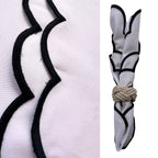 A white fabric napkin with black scalloped trim, shown in a close-up detail view on the left and folded with a rope tie on the right, against a white background.