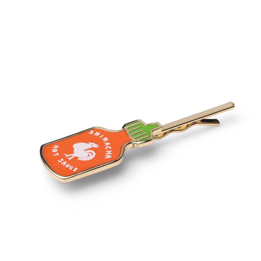 A novelty hair clip designed to look like a Sriracha hot sauce bottle, featuring an orange body with a white rooster logo and text, a green cap, and gold-tone metal edges and clasp.
