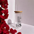 A clear glass tumbler with a bamboo lid and clear straw, printed with 'Thankful Friendsgiving 2026' in black text and leaf graphics. The tumbler is styled on a white surface next to red flowers against a white background.