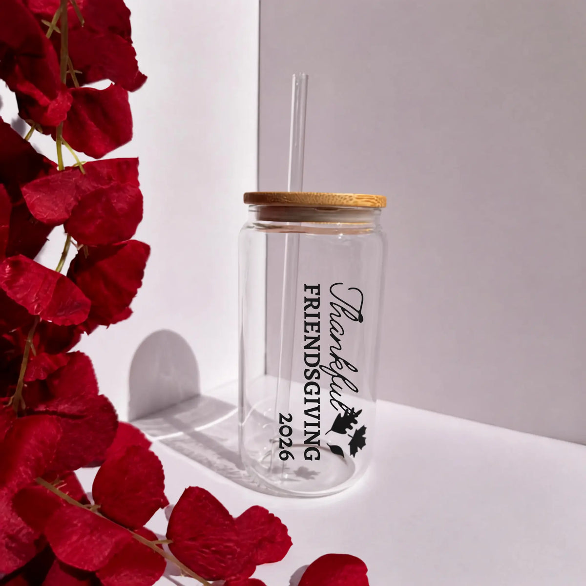 A clear glass tumbler with a bamboo lid and clear straw, printed with 'Thankful Friendsgiving 2026' in black text and leaf graphics. The tumbler is styled on a white surface next to red flowers against a white background.