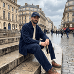 Man sitting on stairs overcast sky wearing a corduroy navy cap with embroidered text "The Friendsgiving Club"