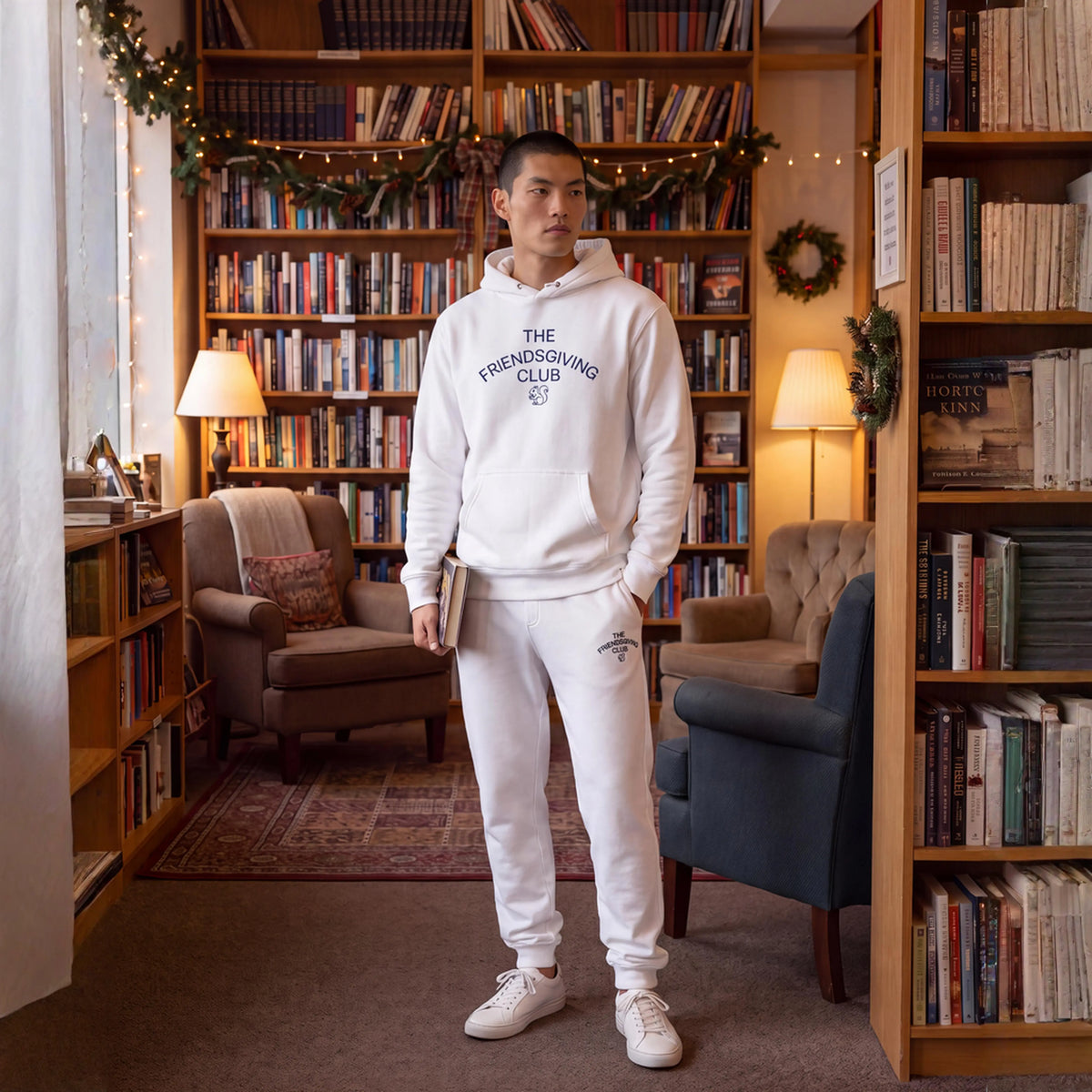 A man wearing a white hoodie and sweatpants set stands in a cozy book-filled room. The hoodie features 'THE FRIENDSLOVING CLUB' printed in navy blue on the chest, and the matching sweatpants have a small logo on the thigh.