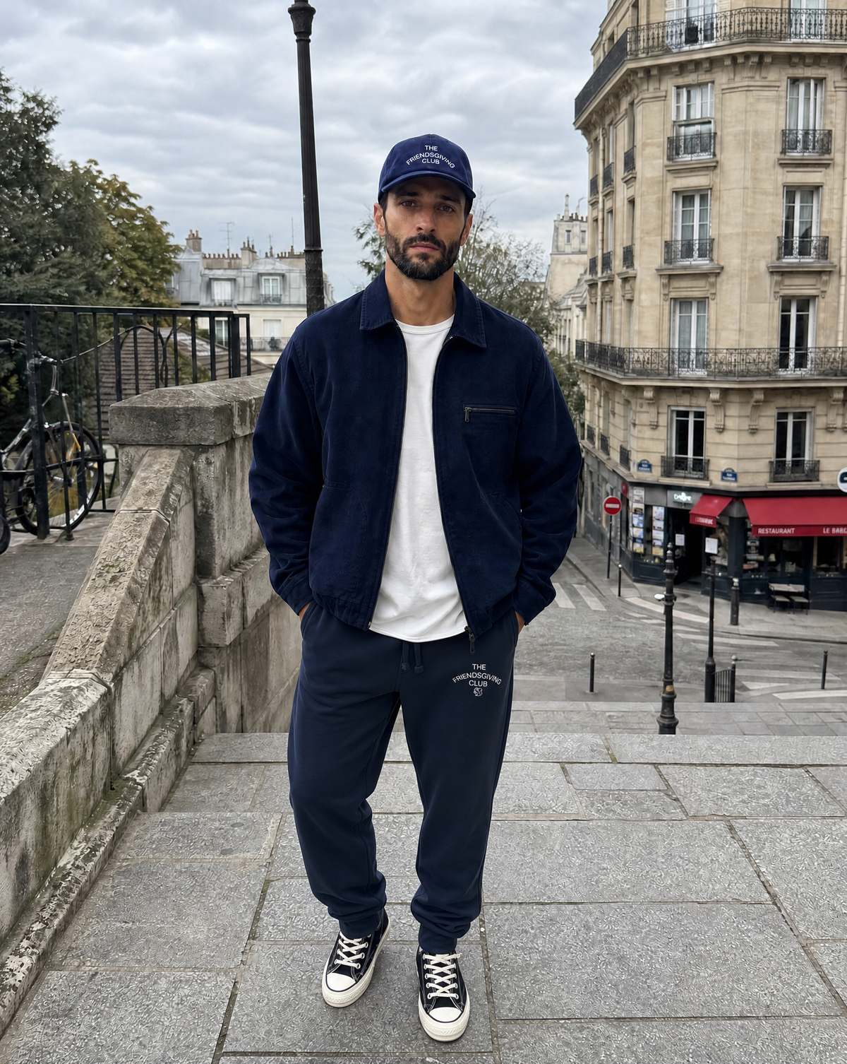 A man wearing a navy blue jacket, matching sweatpants with text The Friendsgiving Club, a navy blue cap, and a white t-shirt stands on stone stairs in an outdoor setting.