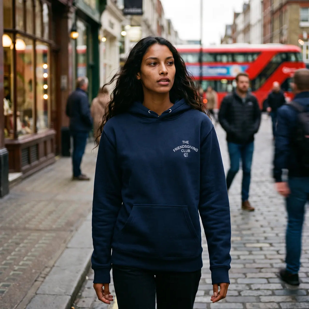 Woman in navy hoodie with white text embroidered "The Friendsgiving Club" walking down cobblestone street
