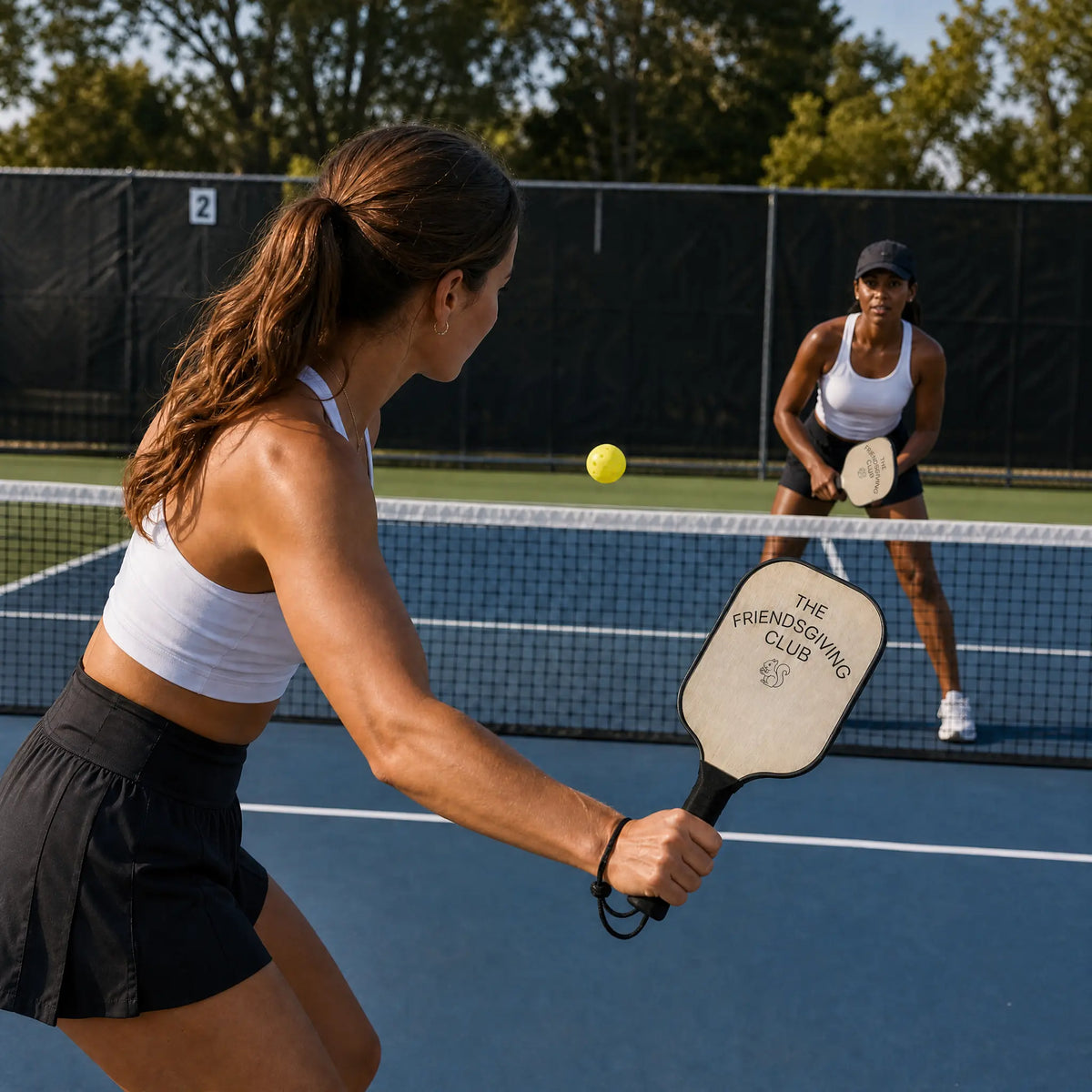 Two women playing pickleball on an outdoor blue court, with one in the foreground holding a paddle that reads 'THE FRENCHGIVING CLUB' and the other waiting for the ball in the background.
