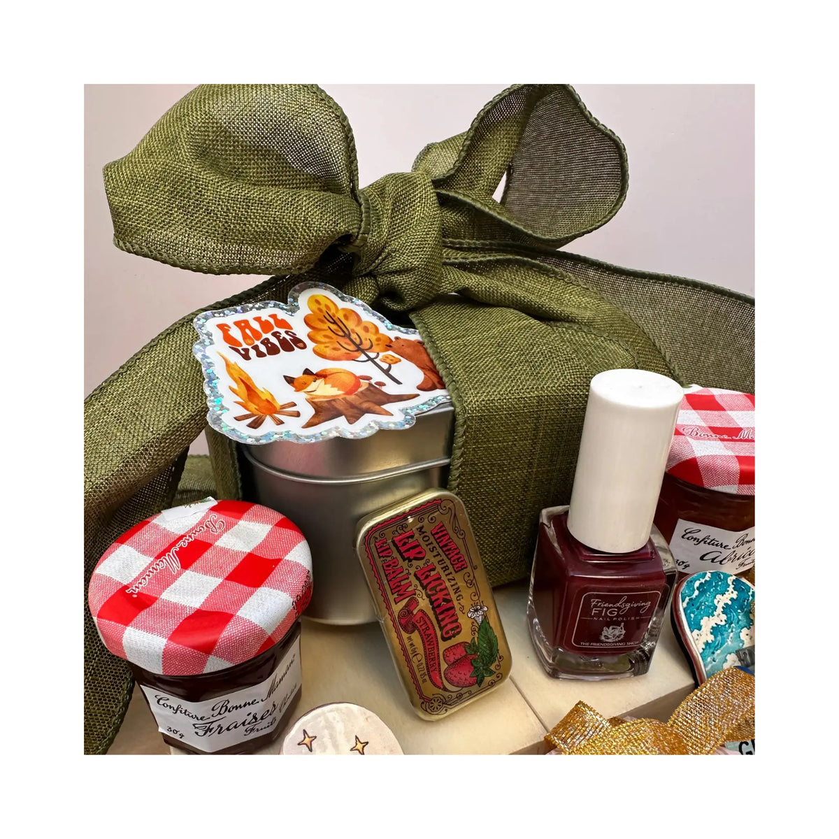 A gift basket arrangement featuring an olive green burlap ribbon bow, a silver tin with autumn leaf artwork, jars with red-and-white gingham lids, a vintage-style candy tin, and a bottle of burgundy nail polish on a light neutral background.