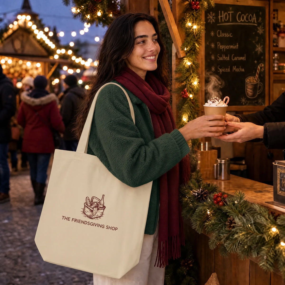 A woman at a festive outdoor hot cocoa stall carries a cream canvas tote bag with a brown logo that reads 'THE FRIENDSGIVING SHOP'. She is wearing a green sweater and a burgundy scarf, smiling as she receives a hot cocoa cup from a vendor.