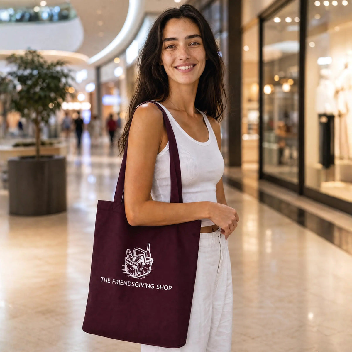 A woman in a white tank top and white pants stands in a shopping mall holding a dark burgundy tote bag with a white graphic and the text 'THE FRIENDSGIVING SHOP'.