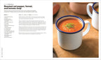 White mug with blue rim filled with roasted red pepper, fennel, and tomato soup on a wooden surface.