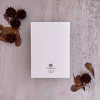 A white rectangular card or invitation with a small black printed crest and cursive text near the bottom, photographed from above on a light textured surface with dried leaves and seed pods arranged around it.