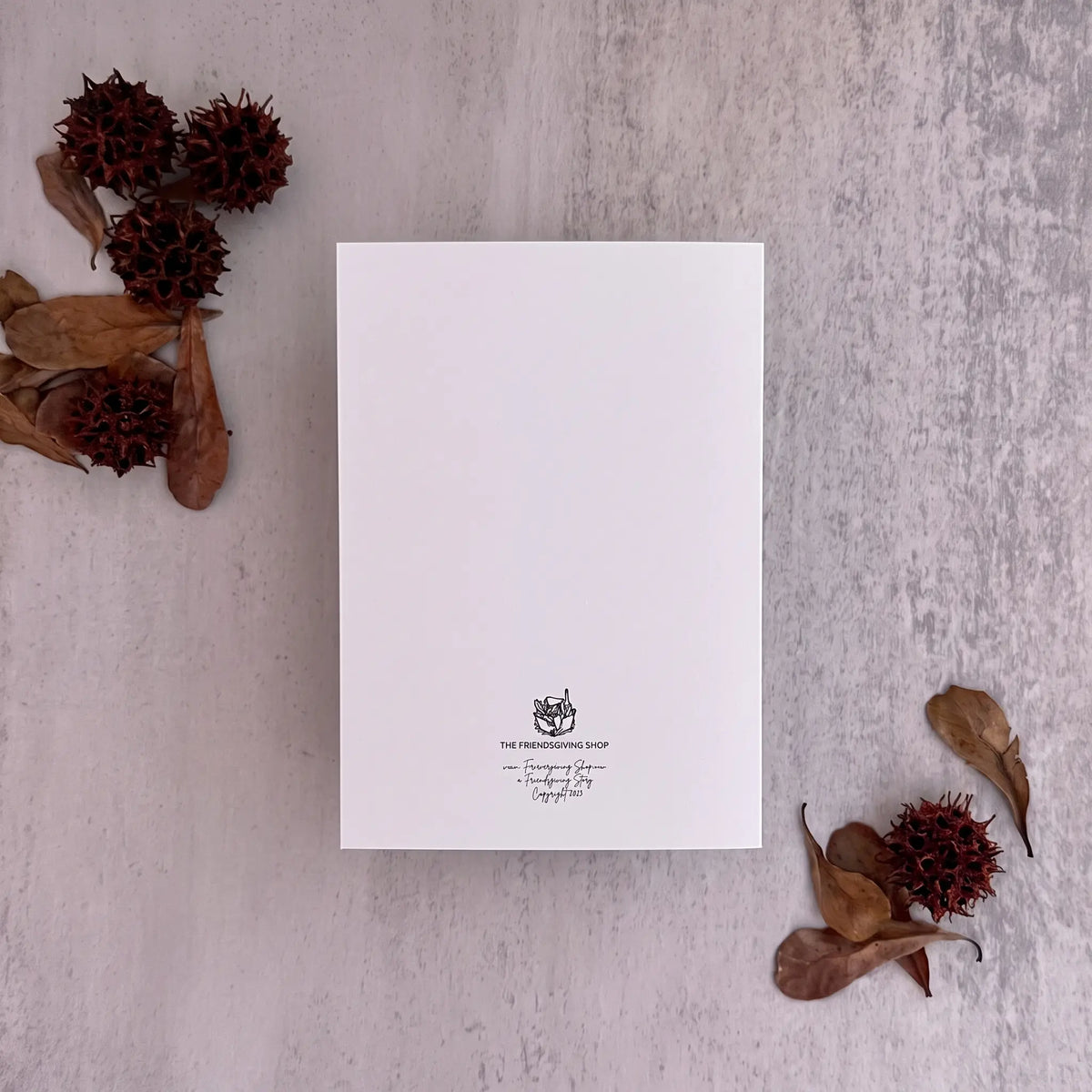 A white rectangular card or invitation with a small black printed crest and cursive text near the bottom, photographed from above on a light textured surface with dried leaves and seed pods arranged around it.