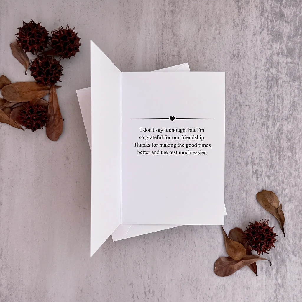 A white folded greeting card with black printed text on the inside, displayed on a light gray textured surface with dried brown leaves and seed pods around it.