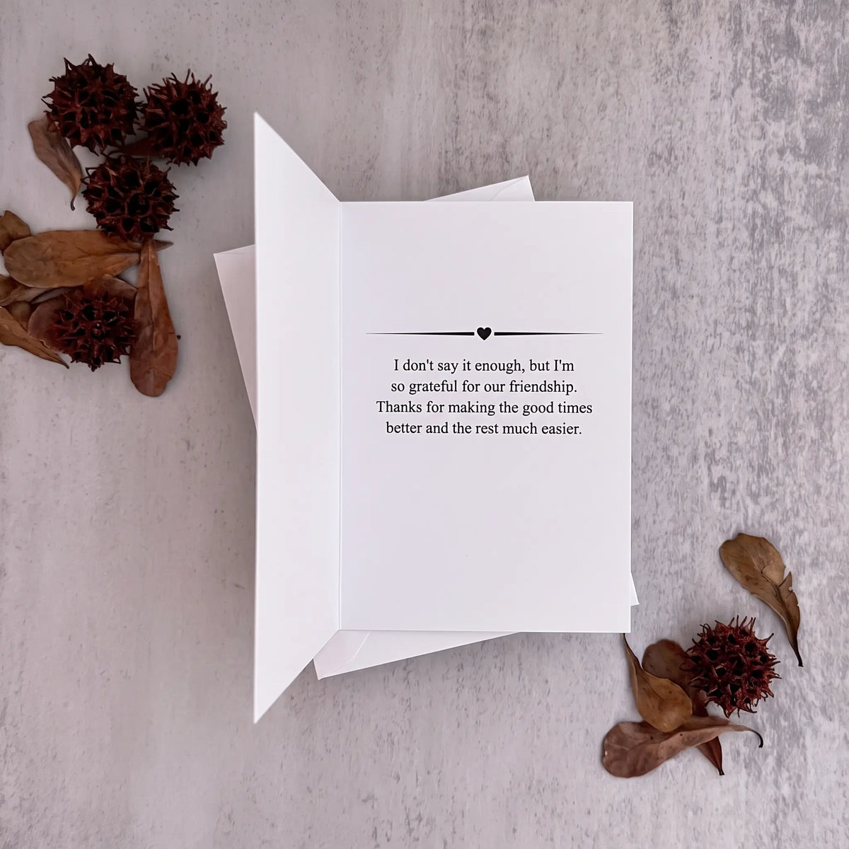 A white folded greeting card with black printed text on the inside, displayed on a light gray textured surface with dried brown leaves and seed pods around it.