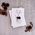 A white greeting card with a black line art illustration of two women and the handwritten text 'To the one who makes everything more fun' with a small red heart, displayed on a textured gray background with dried leaves and tassel flowers.