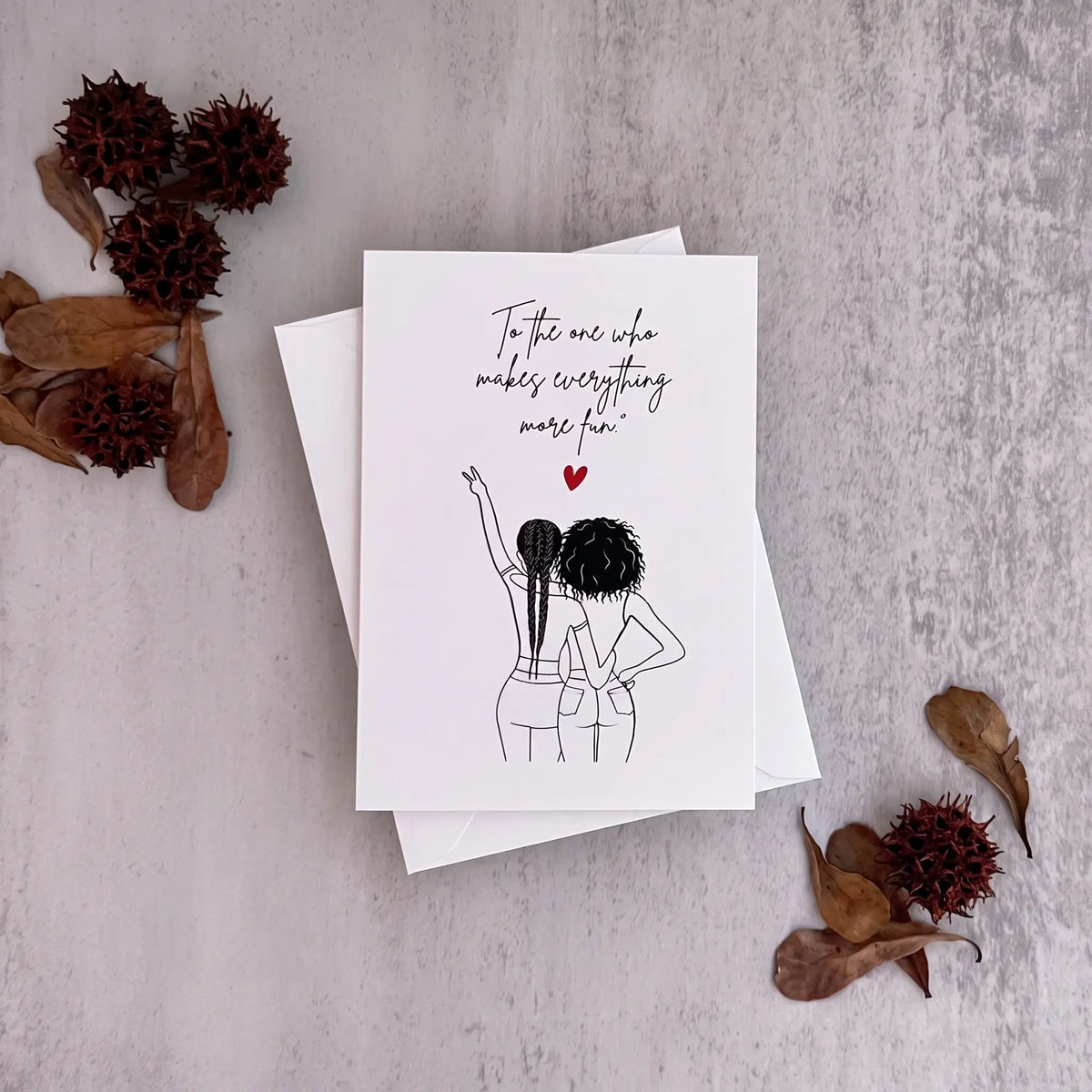 A white greeting card with a black line art illustration of two women and the handwritten text 'To the one who makes everything more fun' with a small red heart, displayed on a textured gray background with dried leaves and tassel flowers.