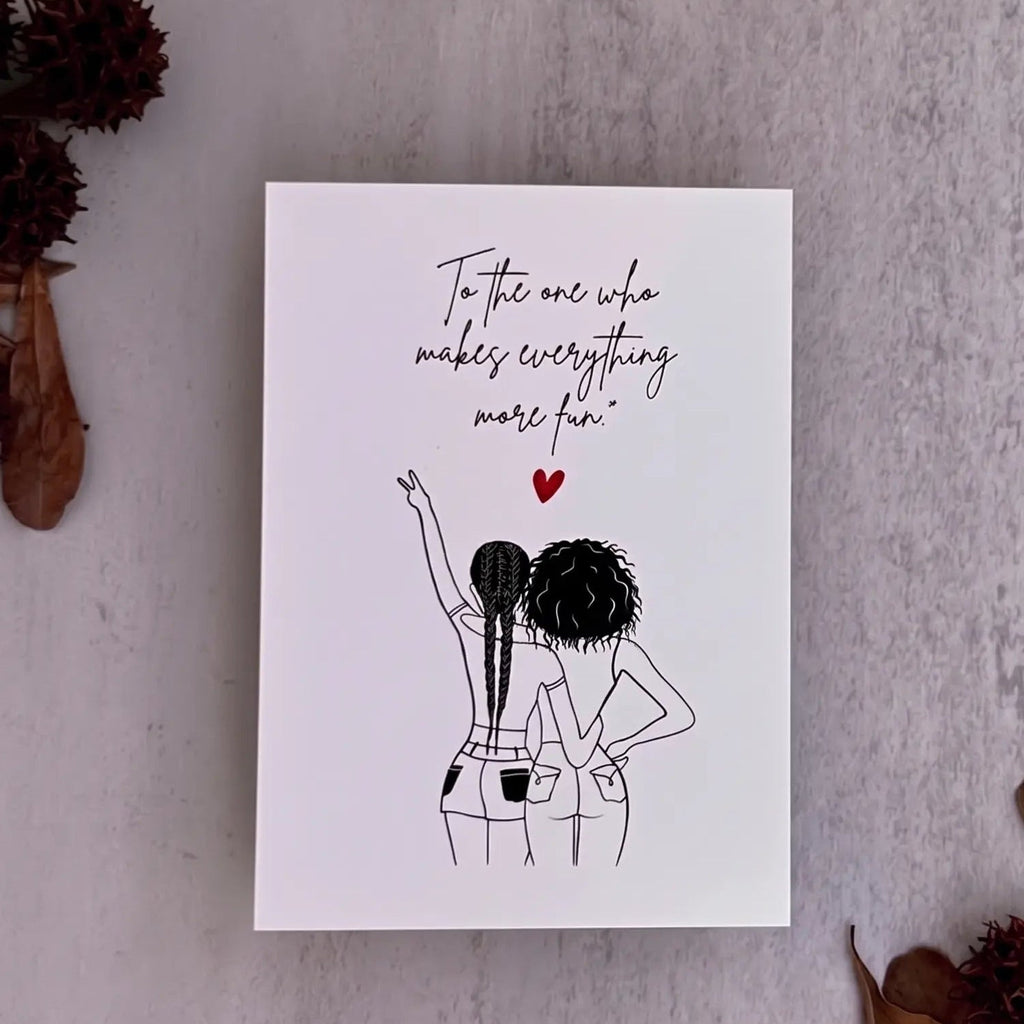 A white greeting card with a black line art illustration of two women from behind and a red heart, placed on a textured gray surface with dried brown leaves and pods around it.