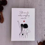 A white greeting card with a black line art illustration of two women from behind and a red heart, placed on a textured gray surface with dried brown leaves and pods around it.
