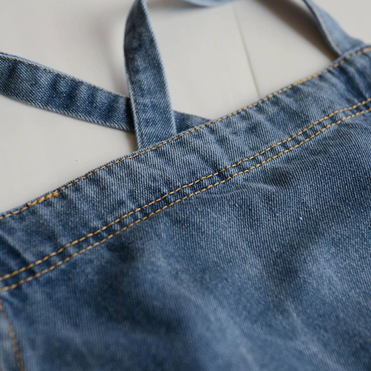 Close-up of a blue denim apron strap and waistband with contrast yellow stitching, laid flat on a light background.