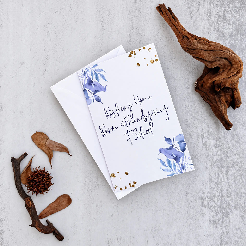 A white Friendsgiving greeting card with blue floral illustrations and gold confetti accents, placed on a light gray textured surface with dried botanical props and a piece of driftwood.