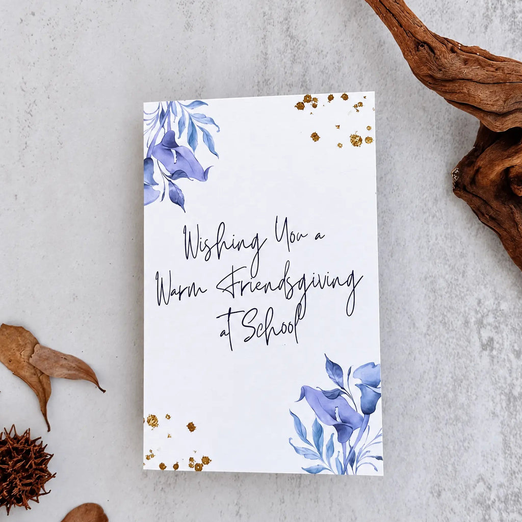 A white greeting card with blue floral illustrations in the corners and gold glitter accents, featuring the handwritten text 'Wishing You a Warm Thanksgiving at School.' The card is photographed in a styled flat-lay on a light gray textured surface with dried leaves, a pine cone, and driftwood around it.