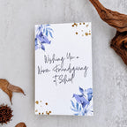 A white greeting card with blue floral illustrations in the corners and gold glitter accents, featuring the handwritten text 'Wishing You a Warm Thanksgiving at School.' The card is photographed in a styled flat-lay on a light gray textured surface with dried leaves, a pine cone, and driftwood around it.