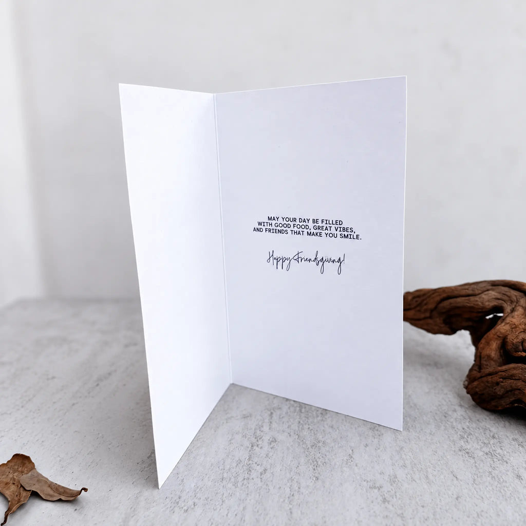 A white folded greeting card standing open on a light stone surface, printed with a short Friendsgiving message and the words 'Happy Friendsgiving' in black. A piece of driftwood sits to the right, and the background is softly blurred.