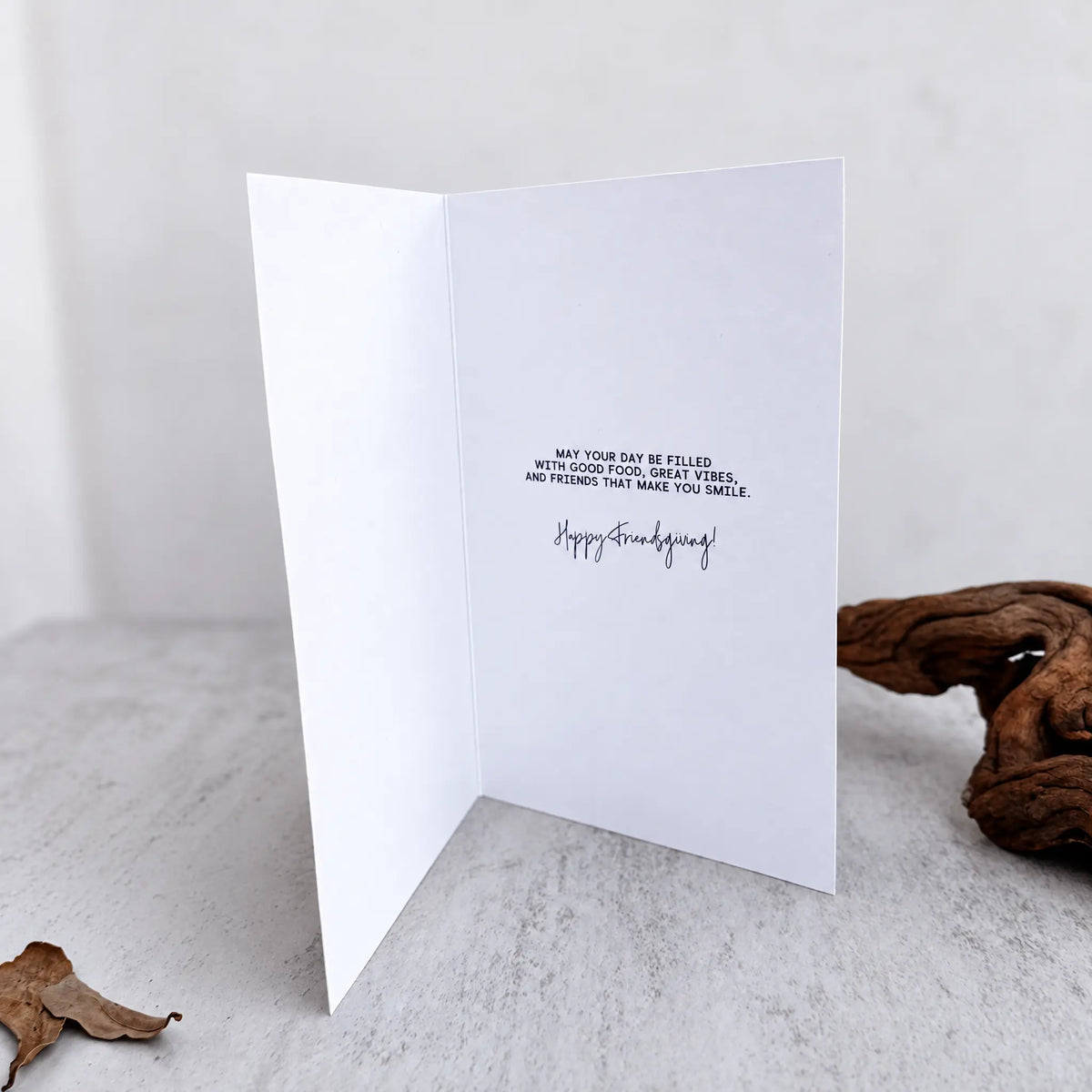 A white folded greeting card standing open on a light stone surface, printed with a short Friendsgiving message and the words 'Happy Friendsgiving' in black. A piece of driftwood sits to the right, and the background is softly blurred.