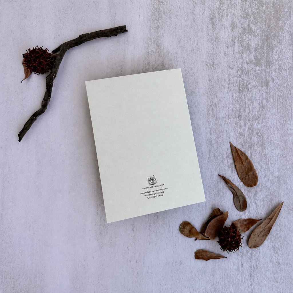 A blank cream-colored letterhead card with a small crest and text at the bottom, styled in a flat-lay with dried leaves and a twig on a textured light gray background.