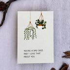 A square white greeting card with a minimalist illustration of two hanging green plants and the text 'YOU'RE A BAD SEED AND I LOVE THAT ABOUT YOU', styled flat-lay on a light textured surface with dried leaves and a small branch.