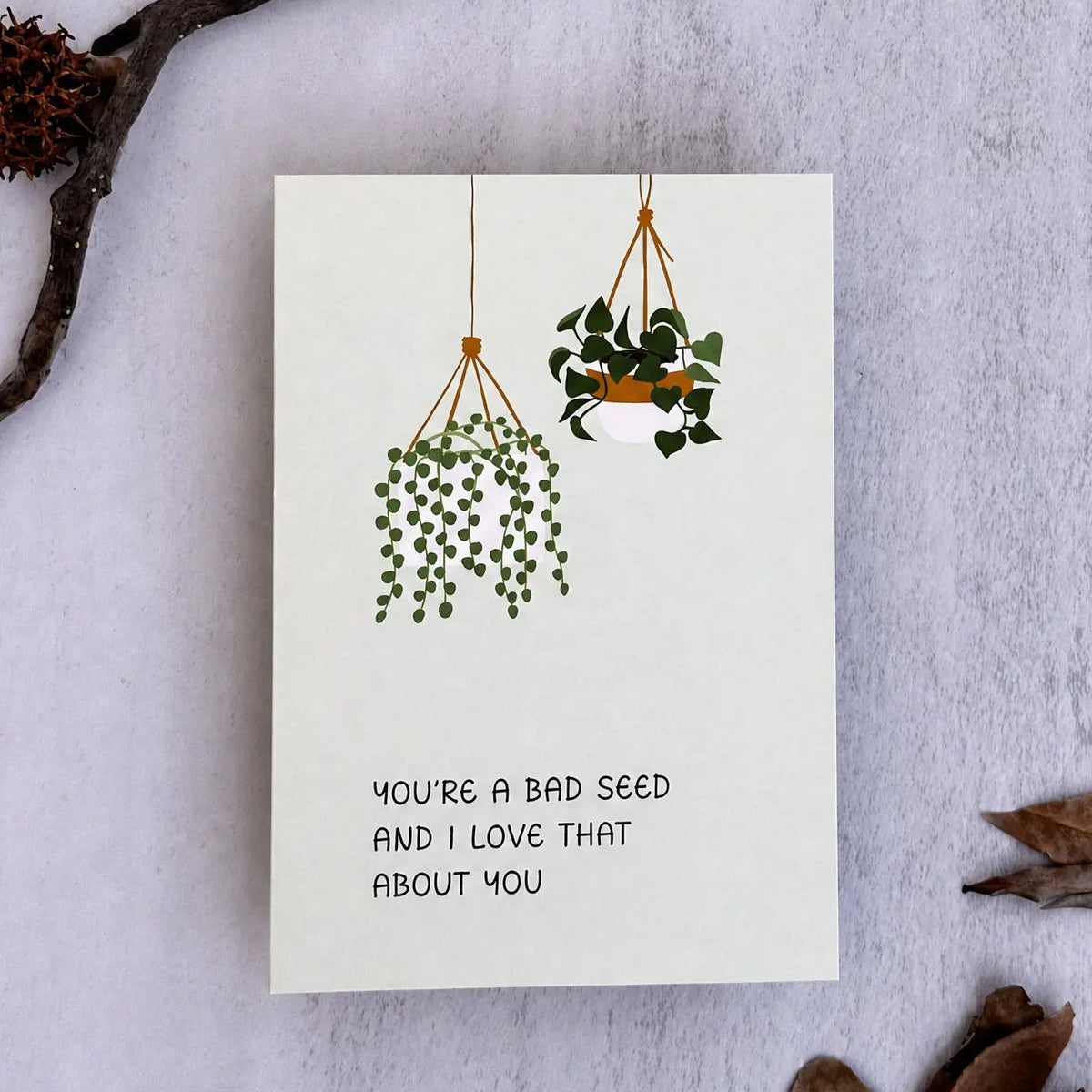 A square white greeting card with a minimalist illustration of two hanging green plants and the text 'YOU'RE A BAD SEED AND I LOVE THAT ABOUT YOU', styled flat-lay on a light textured surface with dried leaves and a small branch.