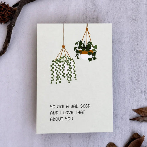A square white greeting card with a minimalist illustration of two hanging green plants and the text 'YOU'RE A BAD SEED AND I LOVE THAT ABOUT YOU', styled flat-lay on a light textured surface with dried leaves and a small branch.