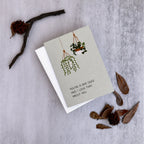 A square greeting card with a light gray-green background, featuring an illustration of two hanging green plants and the printed message 'YOU'RE A BAD SEED AND I LOVE THAT ABOUT YOU'. The card is displayed on top of a white envelope, styled with dried botanical elements on a textured gray surface.