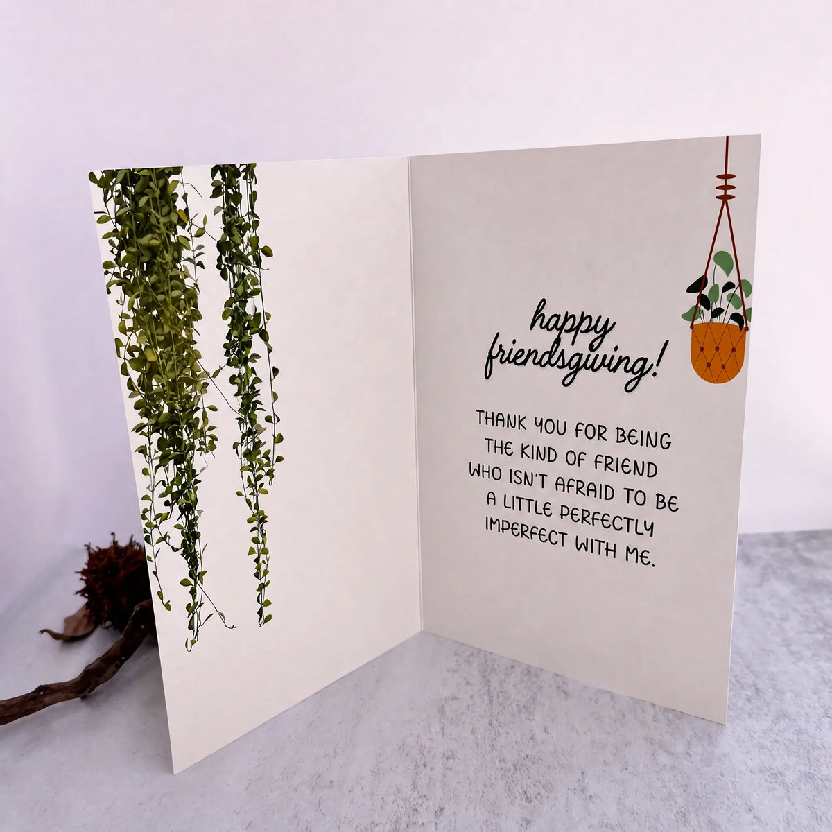 An open greeting card with a warm cream background, featuring green trailing plant illustrations on the left panel and a small potted plant on the right panel with the handwritten-style text 'happy friendsgiving! THANK YOU FOR BEING THE KIND OF FRIEND WHO ISN'T AFRAID TO BE A LITTLE PERFECTLY IMPERFECT WITH ME.' The card is photographed on a light surface against a pale lavender background.
