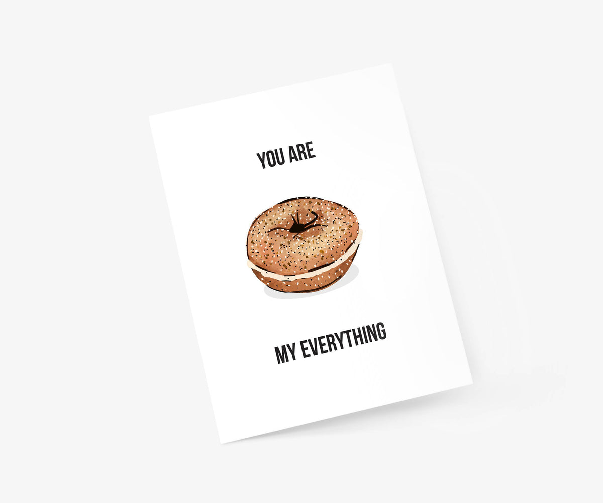 A white square greeting card or print shown at an angle on a light gray background. The card features a watercolor-style bagel with cream filling and the text 'YOU ARE MY EVERYTHING' in black.