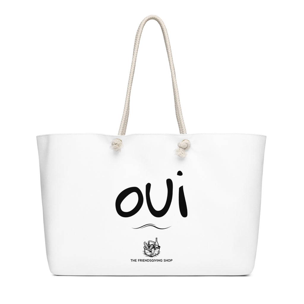 A white canvas tote bag with cream-colored rope handles, featuring the word 'oui' in black script and a small graphic with the text 'THE FRIENDSGIVING SHOP' printed on the front.